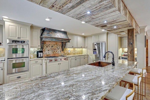 kitchen featuring sink, appliances with stainless steel finishes, a kitchen breakfast bar, light stone countertops, and custom exhaust hood