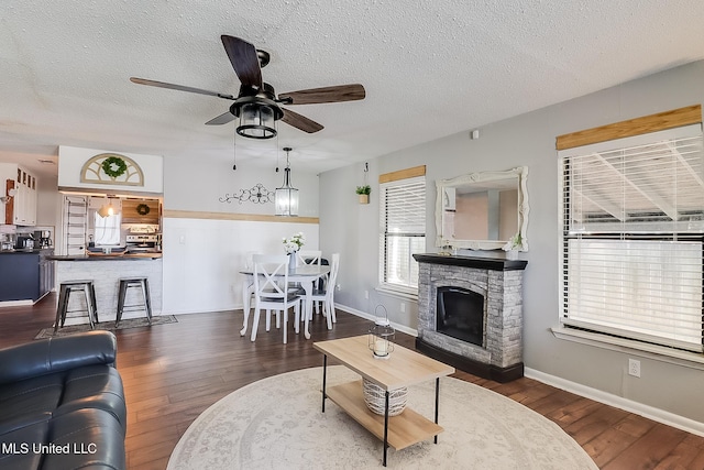 living area featuring a textured ceiling, a stone fireplace, dark wood finished floors, and a ceiling fan