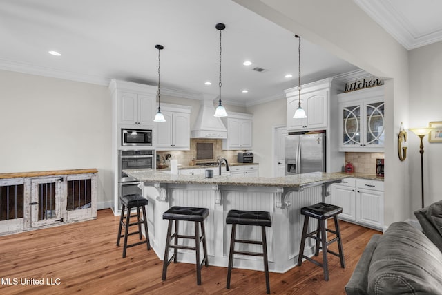 kitchen with appliances with stainless steel finishes, light stone countertops, custom range hood, an island with sink, and white cabinets