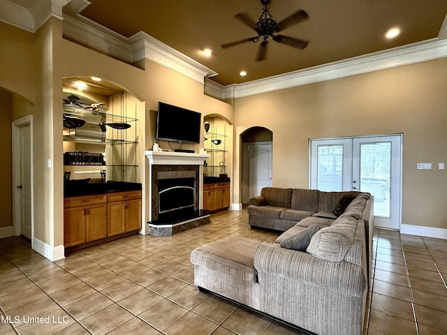 living room with crown molding, a tiled fireplace, light tile patterned flooring, arched walkways, and a ceiling fan