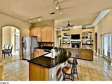 kitchen with stainless steel refrigerator with ice dispenser, dark countertops, arched walkways, a fireplace, and ceiling fan