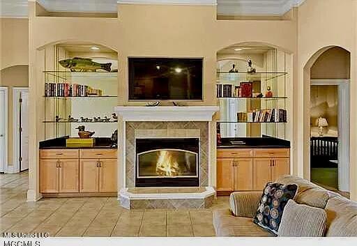 living room with built in shelves, a glass covered fireplace, arched walkways, light tile patterned flooring, and crown molding