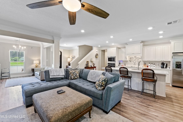 living room featuring decorative columns, ornamental molding, ceiling fan with notable chandelier, light hardwood / wood-style floors, and sink