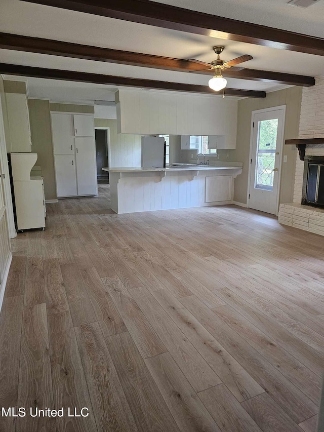 unfurnished living room featuring beam ceiling, ceiling fan, a brick fireplace, and light wood-type flooring