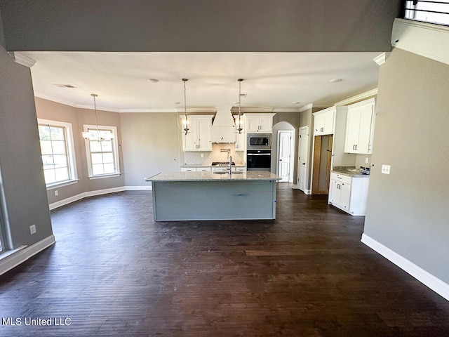 kitchen featuring stainless steel microwave, a center island with sink, oven, hanging light fixtures, and decorative backsplash