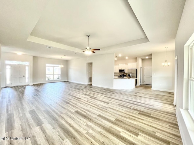 unfurnished living room with baseboards, visible vents, light wood-style floors, ceiling fan with notable chandelier, and a raised ceiling