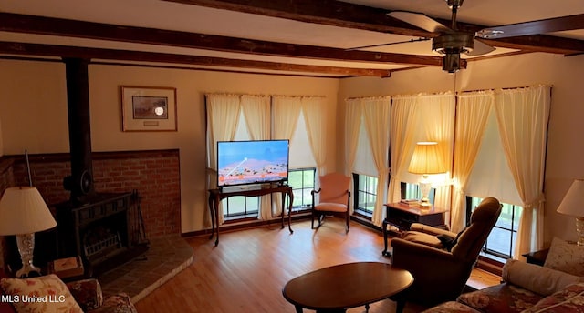 living area with beam ceiling, wood finished floors, a wood stove, and a ceiling fan