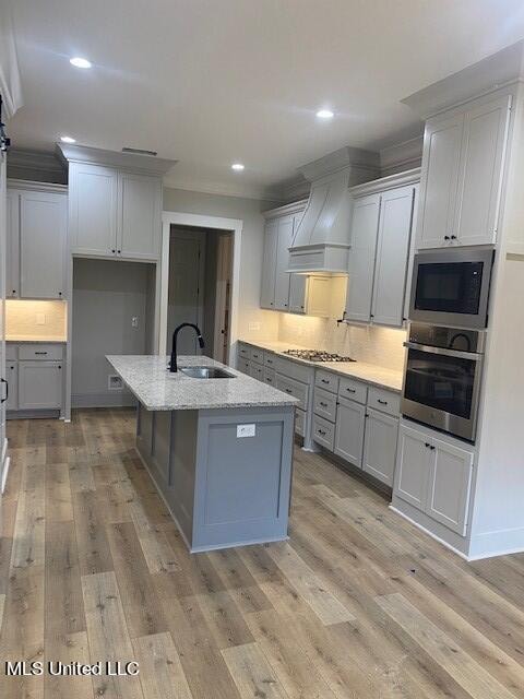 kitchen featuring custom exhaust hood, a center island with sink, appliances with stainless steel finishes, light hardwood / wood-style floors, and sink