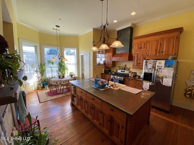 kitchen with refrigerator with ice dispenser, stainless steel gas stove, wall chimney exhaust hood, brown cabinetry, and dark countertops