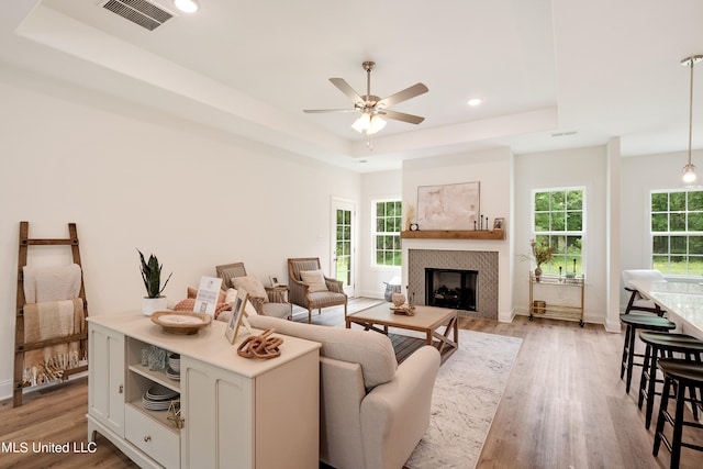 living room with a raised ceiling, ceiling fan, a fireplace, and light wood-type flooring