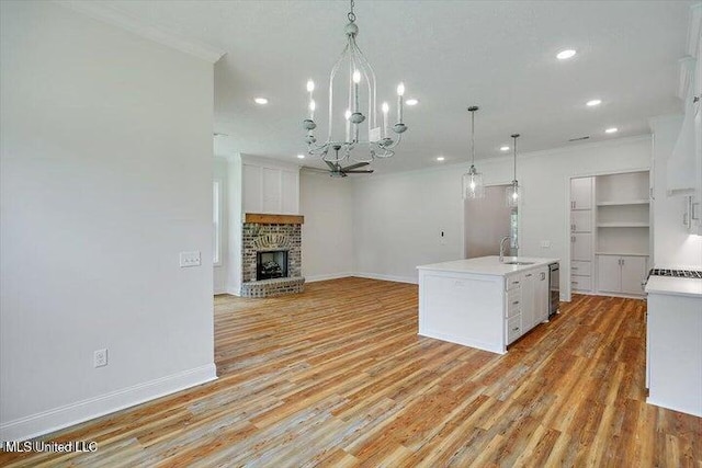 kitchen featuring a stone fireplace, hanging light fixtures, an island with sink, white cabinetry, and light wood-type flooring