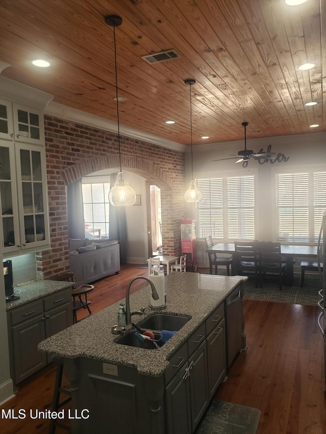 kitchen featuring light stone counters, brick wall, a center island with sink, and sink