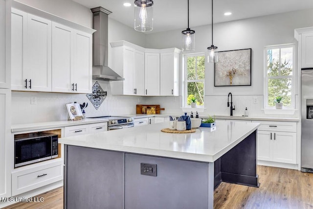 kitchen with wall chimney range hood, white cabinets, decorative light fixtures, a kitchen island, and appliances with stainless steel finishes