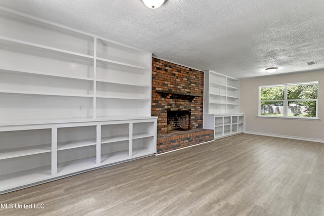 unfurnished living room with a textured ceiling, a brick fireplace, built in shelves, wood finished floors, and baseboards