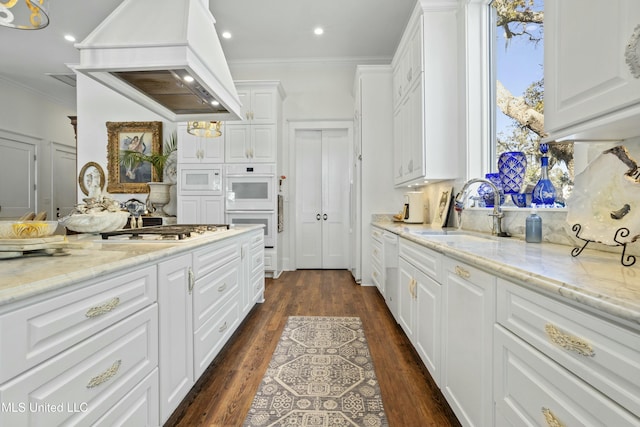 kitchen with white appliances, a sink, white cabinets, custom range hood, and crown molding