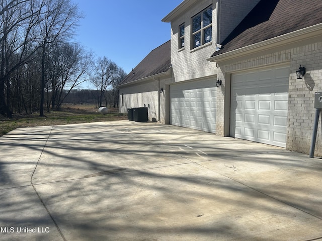 view of property exterior featuring central AC unit, driveway, a shingled roof, a garage, and brick siding