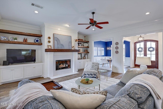 living room featuring light wood-type flooring, a large fireplace, built in features, crown molding, and ceiling fan with notable chandelier