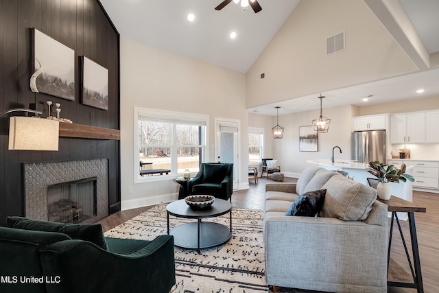 living area with a ceiling fan, high vaulted ceiling, a tiled fireplace, and light wood-style floors