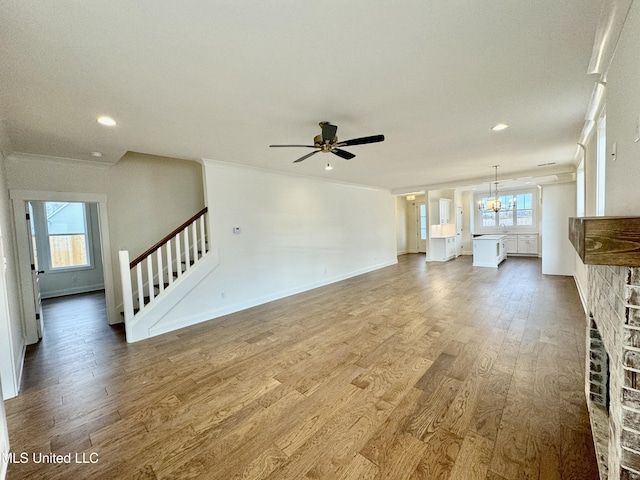 unfurnished living room with a stone fireplace, crown molding, wood-type flooring, and ceiling fan with notable chandelier