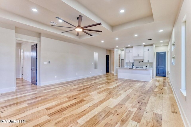 unfurnished living room featuring sink, a raised ceiling, ceiling fan, and light wood-type flooring