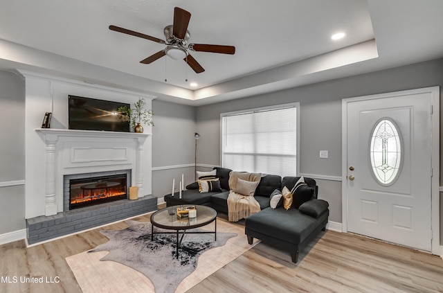 living room with a tray ceiling, a brick fireplace, a ceiling fan, light wood-type flooring, and plenty of natural light