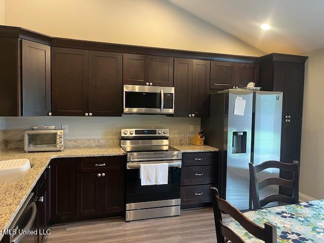 kitchen featuring appliances with stainless steel finishes, lofted ceiling, light wood finished floors, light stone countertops, and dark brown cabinets