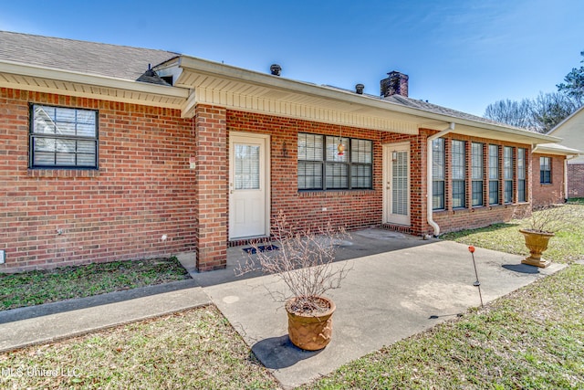 entrance to property featuring a shingled roof, brick siding, and a patio