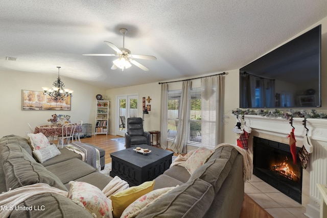 living room featuring ceiling fan with notable chandelier, a textured ceiling, a tile fireplace, and light hardwood / wood-style flooring