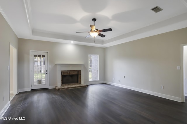 unfurnished living room featuring dark wood-type flooring, ceiling fan, ornamental molding, a brick fireplace, and a raised ceiling