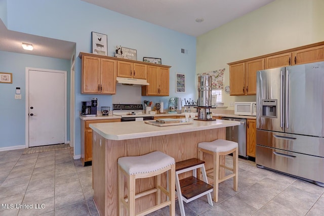 kitchen featuring a towering ceiling, appliances with stainless steel finishes, light countertops, under cabinet range hood, and a breakfast bar area