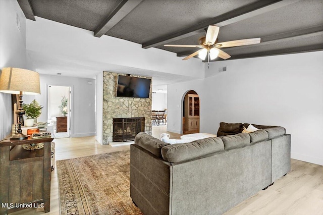 living room featuring a stone fireplace, beamed ceiling, light hardwood / wood-style flooring, a textured ceiling, and ceiling fan