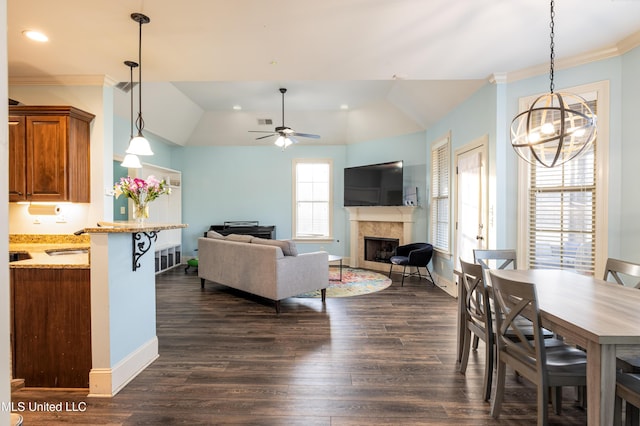 dining area featuring dark wood-style floors, vaulted ceiling, crown molding, a fireplace, and ceiling fan with notable chandelier