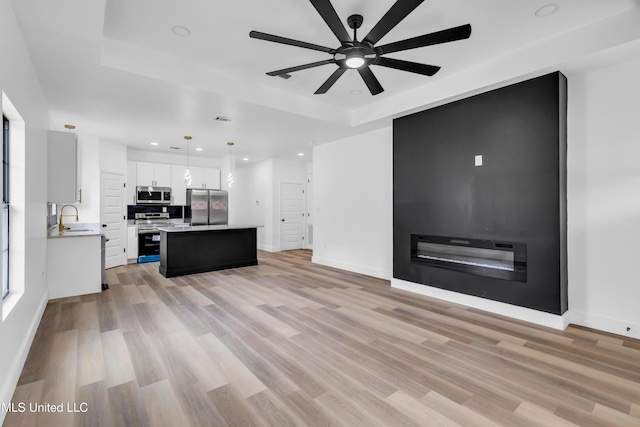 unfurnished living room with light wood-style floors, a sink, baseboards, a ceiling fan, and a tray ceiling