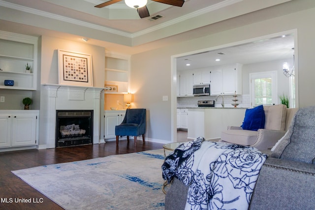 living room with dark wood-style floors, crown molding, a ceiling fan, a fireplace, and built in shelves
