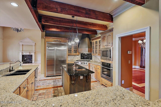 kitchen featuring wall chimney range hood, sink, built in appliances, beamed ceiling, and dark stone counters