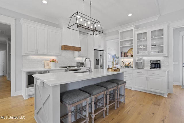 kitchen featuring white cabinets, appliances with stainless steel finishes, glass insert cabinets, a kitchen island with sink, and light countertops