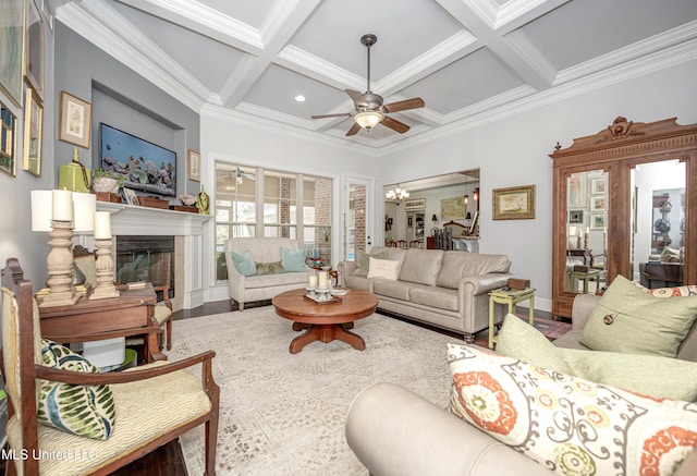 living area featuring beamed ceiling, ornamental molding, ceiling fan with notable chandelier, coffered ceiling, and a glass covered fireplace