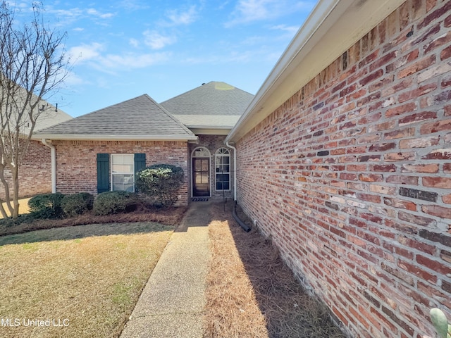 view of exterior entry with a lawn, brick siding, and roof with shingles