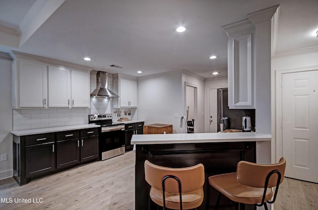 kitchen featuring electric range, wall chimney exhaust hood, white cabinetry, and a breakfast bar area