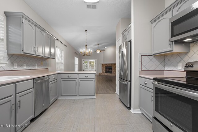 kitchen featuring stainless steel appliances, gray cabinets, a chandelier, a fireplace with raised hearth, and a peninsula