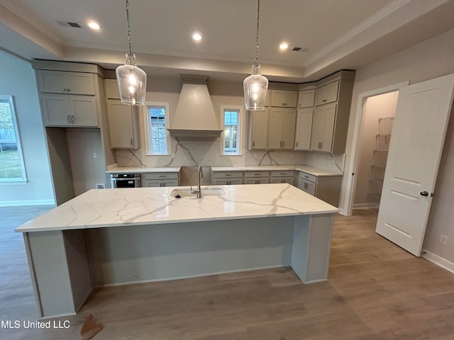 kitchen featuring custom range hood, a kitchen island with sink, sink, and light stone counters