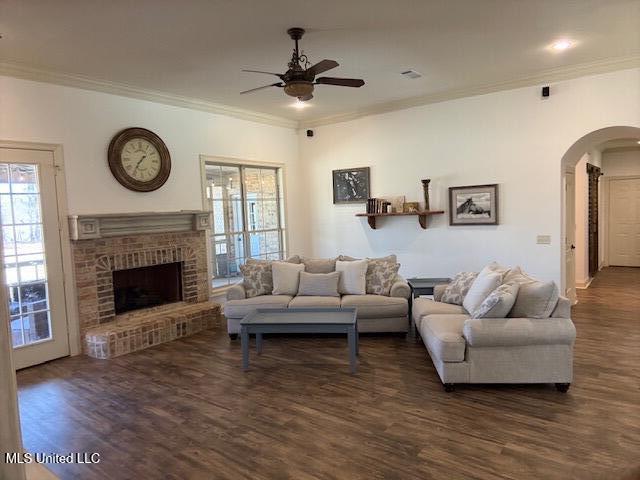 living room featuring dark wood-style floors, arched walkways, a fireplace, crown molding, and a ceiling fan