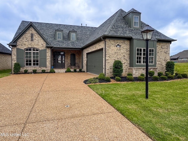 french country home with brick siding, a front lawn, concrete driveway, a garage, and roof with shingles