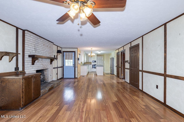 unfurnished living room featuring wood-type flooring, ornamental molding, a brick fireplace, a textured ceiling, and ceiling fan with notable chandelier