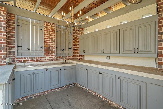 kitchen with beamed ceiling, tile countertops, an inviting chandelier, brick wall, and wooden ceiling