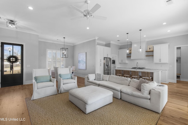 living room with ceiling fan with notable chandelier, ornamental molding, sink, and light hardwood / wood-style flooring