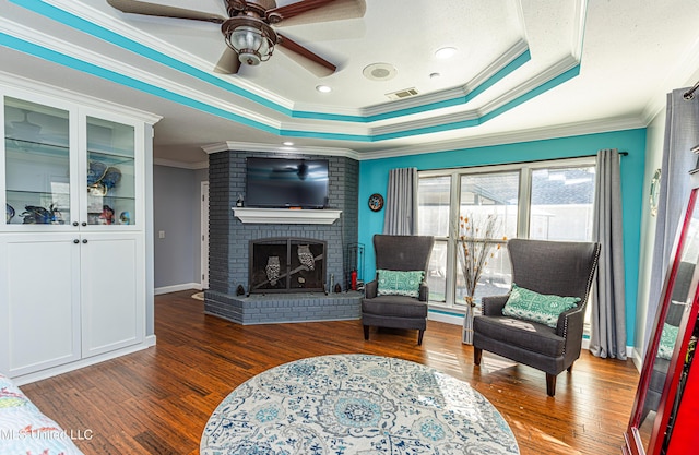 living area with a brick fireplace, crown molding, a tray ceiling, wood finished floors, and a ceiling fan