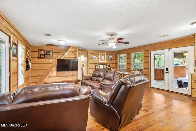 living room featuring french doors, a textured ceiling, wooden walls, light hardwood / wood-style floors, and ceiling fan