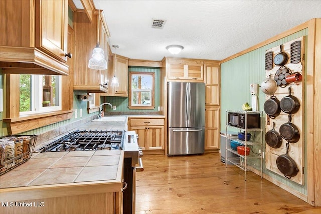 kitchen with tile counters, stainless steel appliances, sink, pendant lighting, and light wood-type flooring