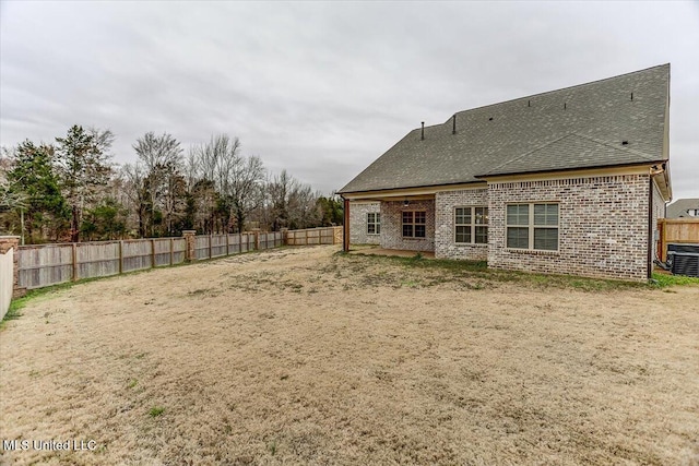 back of property with a fenced backyard, a shingled roof, central AC, and brick siding